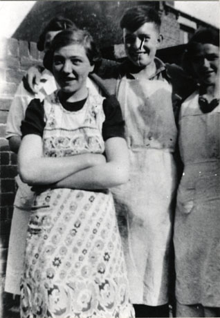 Photograph close-up full-length of a young woman wearing a pinafore; behind her are two other women wearing aprons and a young boy also wearing an apron; they are standing in front of the wall of the backyard of a house. the photograph is described as Mr. Duncan Semens and Staff, Easington Colliery