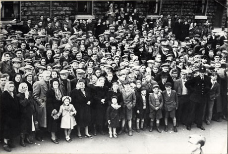 Photograph of a group of approximately one hundred people standing near the walls of houses, described as Group at Pit Head, 1951 (after Easington Disaster)