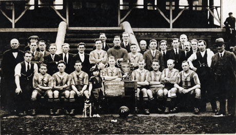 Photograph of the eleven members of the Easington Colliery Welfare Football Team, with nineteen other men, a child dressed in a jester's costume, and a dog; the photograph is taken in front of steps leading up to a stand; in front of the players is a table with a notice reading: Easington Colliery Welfare League Champions 1929-1930 Durham Hospital Cup Winners Finalist National Orphanage Cup 1930
