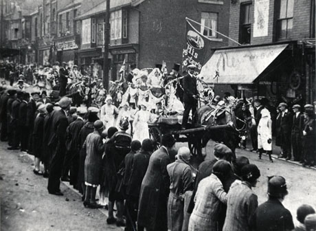 Photograph showing a float progressing along a road, followed by other floats, watched by spectators lining the route; the facades of the shops on the street can be seen and the name Moore's Stores identified; an advertisement for His Master's Voice can also be seen; the photograph is identified as Easington Colliery Carnival