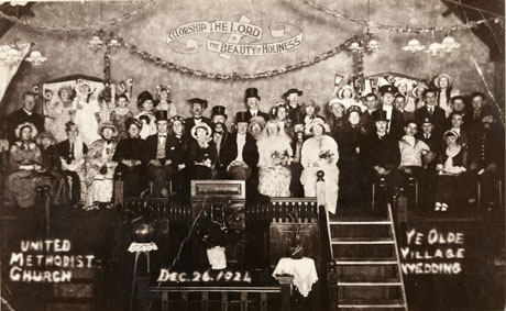 Photograph showing forty people posed on a stage in nineteenth century costume for a production of The Olde Village Wedding in United Methodist Church; behind the cast are the words Worship the Lord In the Beauty of Holiness inscribed on the wall; the photograph bears the date Dec, 26 1924; the photograph has been identified as being in Easington Colliery