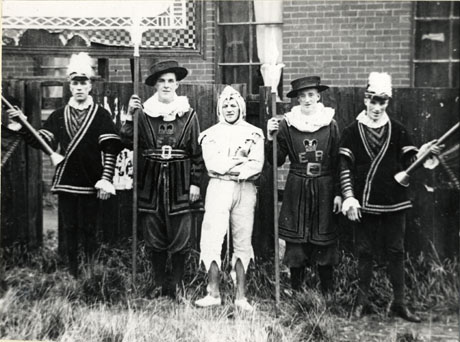 Photograph of four men in fancy dress photographed against a fence; two men are holding the poles of a banner which is out of shot; two men are dressed as beefeaters, two as heralds, and one as a jester; they are described as taking part in the Carnival at Easington Colliery in 1932