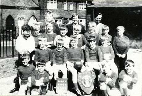 Photograph of a group of seventeen boys aged approximately ten years, described as the Easington Colliery Junior Mixed School Football Team, with three men; in front of the boys are two trophies