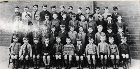 Photograph of a group of forty seven boys aged approximately six years photographed against the wall of a brick-built building; a child on the front row is holding a notice reading Easington Colliery Junior Boys' School Class 1 1929