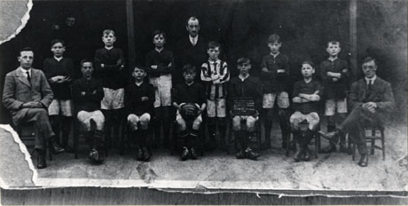Photograph of the eleven boys of Easington Colliery Boys' Football team,with three men; a boy is holding a football identifying the team and another is holding an illegible notice giving details of the trophies won by the team