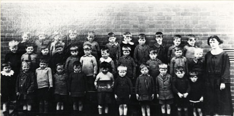 Photograph of thirty three boys in an infants' class at Easington Colliery School; the children are posed against a brick wall and are accompanied by their female teacher; the boy fourth from the left on the front row has been identified as Ted Leadbitter, formerly Member of Parliament for Hartlepool, and the fifth from the left on the front row has been identified as Mr. Rowcroft