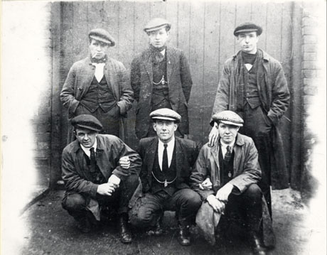 Photograph of six men in suits and flat caps posed against a high gate set in a wall; three are standing and are three are crouching; the men are described as miners of Easington Colliery; they appear to be in their twenties or thirties