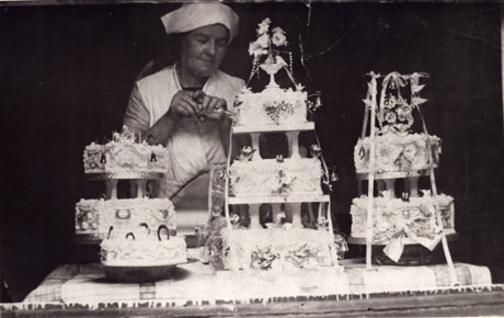 Photograph of a woman in cap and overall standing behind two three-tiered wedding cakes and one two- tiered one all of which are elaborately iced;the woman identified as Mrs. Holmes is piping icing on to the wedding cake in the centre of the photograph