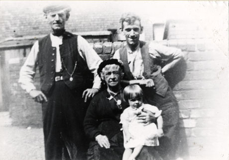 Photograph of two men, a woman, and a child posed against a wall behind a house; one of the men is wearing an open waistcoat with a watch chain; the second man who appears to be younger is also wearing an open waistcoat; the woman, who is seated in front of the men, is elderly and wearing a long dark dress; the child who is sitting on the lap of the woman is aged approximately four years; the group has been identified as four generations of the Humphrey Family