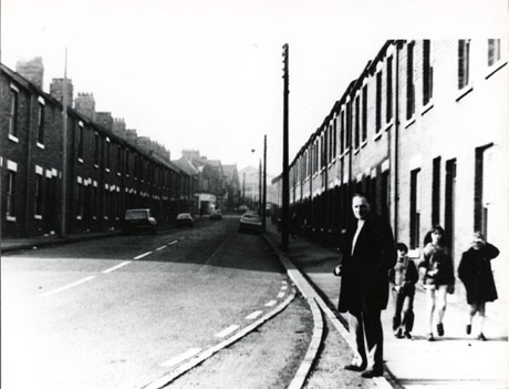 Photograph described as Corner of Colliery Offices, Easington Colliery, showing a street with terraced houses on both sides receding into the distance; a number of cars are parked in the street; a man wearing an overcoat is standing on the pavement in the right-hand side of the photograph; behind him three children can be seen walking towards the camera