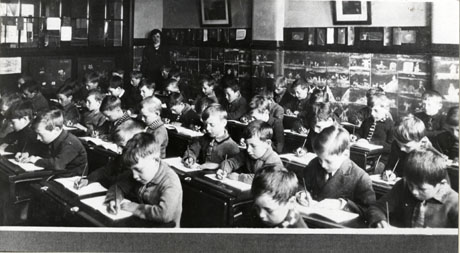 Photograph of the interior of a classroom at Easington Colliery Junior Mixed School showing approximately forty boys aged approximately eight years seated at desks in rows writing with pencils in exercise books; there are drawings on the walls and a schoolteacher is standing in the corner of the classroom