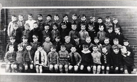 Photograph of a group of forty nine boys, aged approximately seven years, posed against a brick wall; a boy in the front row is holding a notice which reads Easington Colliery Junior Boys. Class 4a 1927