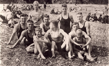 Photograph of a group of seven young men a boy and a girl in swimming costume on a beach, showing groups of other people sitting behind them on the beach; the beach appears to consist of shingle