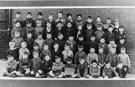 Photograph of a group of fifty four boys, aged approximately seven years, posed against a brick wall and a drainpipe; a boy on the front row is holding a notice which reads Easington Colliery Junior Boys. Class 1b. 1927