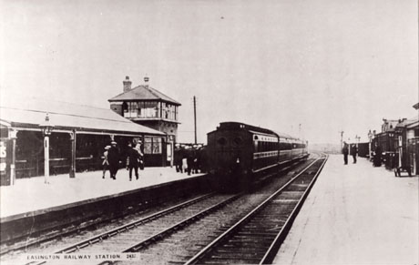 Postcard photograph entitled Easington Railway Station. 2437, showing the tracks, both platforms, the signal box, the rear of a train of coaches drawn up at one platform with a group of indistinct people waiting to board the train; three indistinct figures can be seen on the opposite platform
