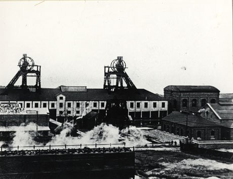 Photograph of the exterior of the buildings of Easington Colliery, showing the buildings in the middle distance with an industrial process generating steam taking place in the foreground; two or three small indistinct figures can be see to the right of the photograph