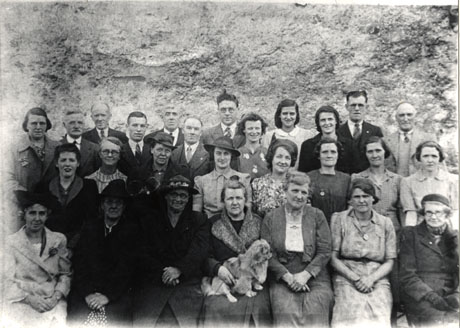 Photograph of a group of eight men and nineteen women posed against a stone wall or a cliff, described as the members of an outing from St, John's Methodist Church, Easington Colliery; a woman on the front row is holding a small dog