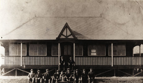 Photograph of the exterior of a pavilion, showing the facade of a one-storied building with a veranda and six large windows; ten men, one girl, and a dog can be seen sitting on the steps in front of the pavilion; the pavilion is described as the Welfare Pavilion, Easington Colliery