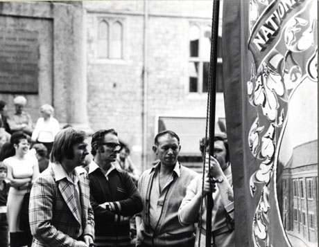 Photograph of four men standing with the Easington Colliery banner, with groups of people behind them and with part of the plinth of the statue of the third Marquess of Londonderry and the wall of the Markets, Durham City, behind them; very little of the banner can be seen