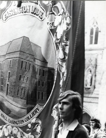 Photograph of the banner of Easington Lodge, showing a picture of the Welfare Hall, Easington Colliery on the banner; the banner is being carried, during the Miners' Gala, by a young man, behind whom the west wall of St. Nicholas's Church, Durham City can be seen