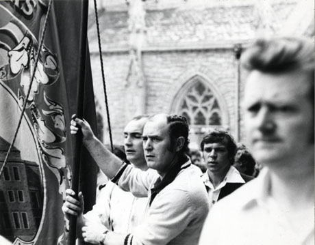 Photograph of four men holding the Easington Lodge banner during the Miners' Gala in The Market Place, Durham City; the exterior of part of the church of St. Nicholas can be seen behind the men; little can be seen of the banner