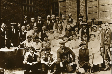 Photograph of the staff of a soup kitchen consisting of twenty five men and twelve women posed against the outside wall of a brick-built building, with a large copper, buckets, ladles and a bowl; the photograph is described as Easington Soup Kitchens, 1921