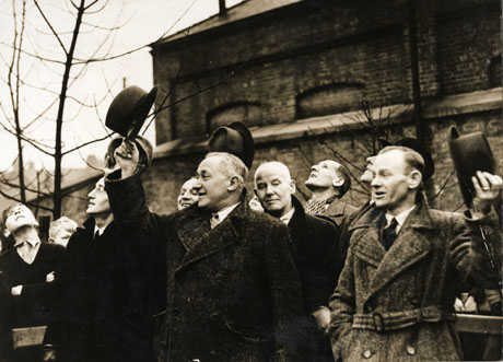 Photograph showing Emmanuel Shinwell in the centre of the photograph accompanied six other men and watched by two boys with the exterior of, possibly colliery, buildings in the background; the men, including Shinwell, are raising their hats in celebration; the photograph is described as marking Shinwell's visit on Vesting Day (the nationalisation of the coal industry) on 1 January 1947