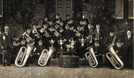Photograph of the members of the Easington Colliery Welfare Band in uniform and with their instruments, posed outside a large brick-built building; in front of the band are five trophies and, to either side, are two men in ordinary dress