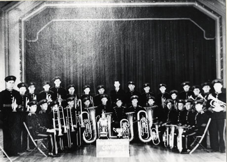 Photograph of the members of Easington Colliery Welfare Band posed on a stage in a theatre or hall; the members are in uniform and with their instruments; in front of them are two trophies and a nearly illegible notice indicating that the band was Durham County Champion in 1950-1951 and in 1951-1952