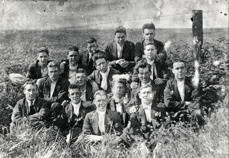Photograph of a group of sixteen young men sitting in the grass in a field in front of a barbed wire fence, with indistinct farmland behind them; they are described as Group of Strikers 1926