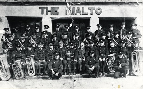 Photograph of the members of the Easington Colliery Welfare Band, posed in uniform and with their instruments outside The Rialto; in the middle of the front row is a man in ordinary clothes