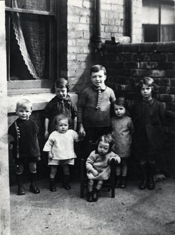 Photograph of seven small children posed in the backyard of a house in the angle where the wall dividing the yard from its neighbour meets the wall of the house; a net curtain can be seen in the window of the house; the children range in age from approximately ten years of age to approximately eighteen months; they are described as being members of the Roberts Family as Children; Station Road, Easington Colliery
