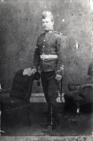 Photograph of a man in soldier's uniform posed in a photographer's studio; the soldier is wearing a tunic with frogging, a forage cap and carries a baton ; he is standing beside a chaise longue and is resting his hands in its back beside his discarded gloves; he is identified as  Mr. Parkin- First Under Manager