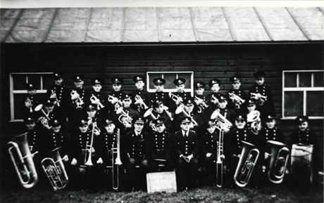 Photograph of the members of Easington Auxiliary Fire Service Band posed outside a low wooden building in uniform and with their instruments; one of the members has been identified as Mr. Peacock in the centre
