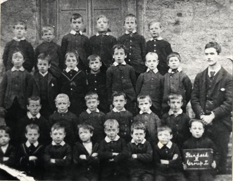 Photograph showing twenty seven boys, aged approximately eight years, posed against the wall of a building in four rows; a man is standing on the right; a boy on the front row is holding a notice reading: Standard V Group I; they have been identified as being pupils at Easington Junior School