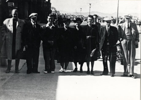 Photograph of a group of five men and four women standing in a line across a pavement with crowds of people in the background and, behind them, the vague outline of hills; the photograph is described as Good Templars Outing in Wales;the atmosphere of the photograph certainly appears to be that of a holiday resort