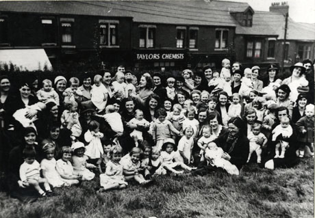 Photograph of a group of approximately forty women with babies and small children, sitting on grass in Seaside Lane with a row of houses and shops behind them; the name Taylors Chemists can be seen