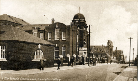 Postcard photograph entitled The Schools, Seaside Lane, Easington Colliery showing the exterior of the schools from the opposite side of the road; the front and side of the nearer building can be seen and the side of the building further down Seaside Lane; a number of passers by can be see on the pavement