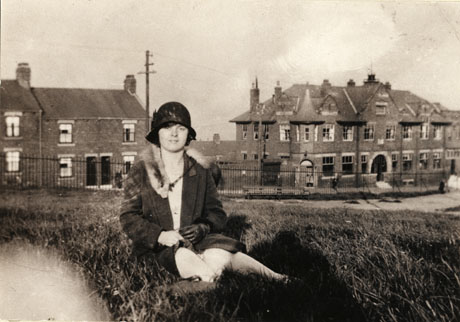 Photograph of a young woman sitting on grass in, possibly, a park, with houses and a large building, identified as The Trust Hotel, on the opposite side of the road; the woman is wearing a cloche hat, an overcoat, and a fur tippet; she is also wearing leather gloves
