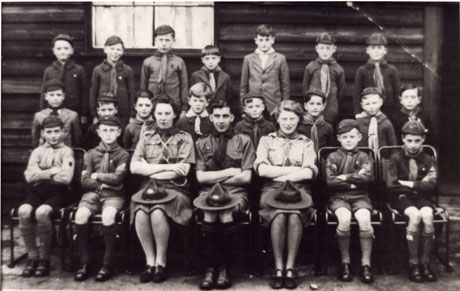 Photograph of a group of nineteen cub scouts with their two female leaders and one male leader, posed outside a building constructed of corrugated iron; all but one of the cubs are in uniform, as are the leaders