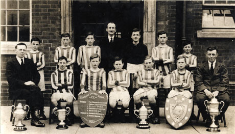 Photograph of the eleven members of the Easington Colliery School Football team, with three men, posed outside the entrance of an imposing brick building; the footballers are in their kit and there are five trophies on the ground in front of them; a boy on the front row is holding a shield detailing their successes, as follows: Winners-East Durham Schools' League; Durham County Hospital Cup; B&O Shield; North Durham and Durham Cup;Bethune Cup. Finalists-Durham County Schools Cup; Horden Hospital Cup.