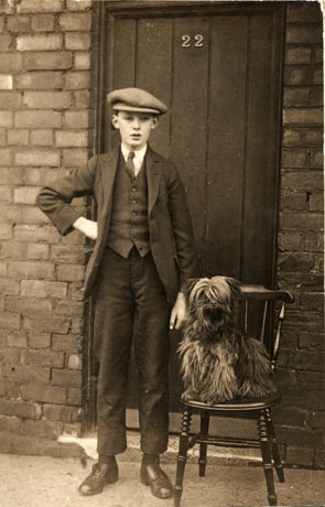 Photograph of a young boy identified as George A. Sutherland, aged 15 years, posed outside a door bearing the number 22 and described as being in Brown Street, Easington Colliery; the boy is wearing a suit with a waistcoat, collar and tie, and cap; he is standing beside a chair on which sits a small long-haired dog