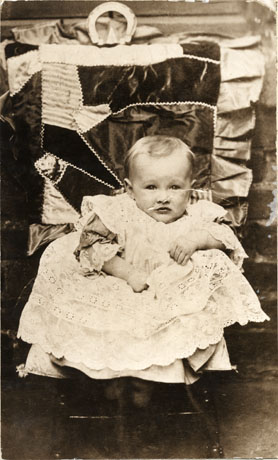 Photograph of a baby dressed in a lacy pinafore and propped on a chair with cushions behind him; he is identified as Robert Smith Mills of Easington Colliery