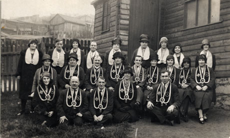 Photograph of seven men and sixteen women posed outside a wooden building with further wooden buildings in the distance; each member of the group is wearing a collar round his or her neck bearing insignia which is identified as that of The Good Templars