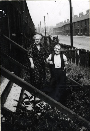 photograph of an elderly man and woman standing on the steps of a building, most likely their house; behind them the rest of the street can be seen with terraced houses on both sides; the street has ben identified as Station Road, Easington Colliery