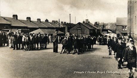 Postcard photograph entitled Schools (Senior Boys), Easington Colliery showing the playground of the school surrounded by houses and other buildings; groups of boys are in the playground drawn up their form groups in preparation for returning to the classroom; a schoolmaster is standing with each group; on the right of the photograph a group is walking into school