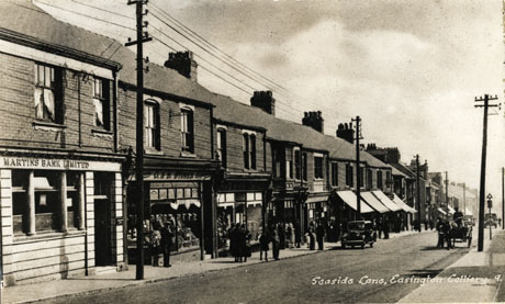 Postcard photograph entitled Seaside Lane, Easington Colliery 9 looking down the street from the right-hand side; a row of shops can be seen; the only names that can be read are Martins Bank Limited, and G & S Stores, a greengrocer; a number of indistinct people are on the pavement and three cars and a cart can be seen