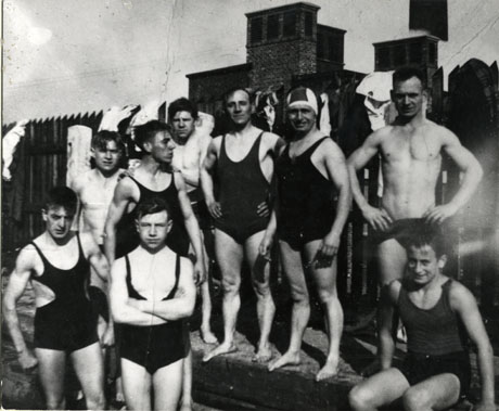 Photograph of nine young men in swimming costume photographed in front of a fence with the top of brick building showing over the fence; the young men are all standing up and are described as members of the Easington Swimming Club