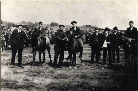 Photograph , described as Pit Pony Racing, Easington Colliery, Welfare Park, 1926, showing, in the foreground, three ponies with boys sitting on them and a man standing at the head at each of them; a group of three men is standing near the ponies; in the background a crowd of people can be seen, and behind them a slope with huts on it