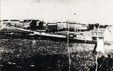 Photograph showing, in the middle distance, a road with fields on one side and four intermittently spaced houses and shops on the other; in the distance a chapel can be seen and a row of houses; the photograph is described as Seaside Lane, Easington Colliery, 1910